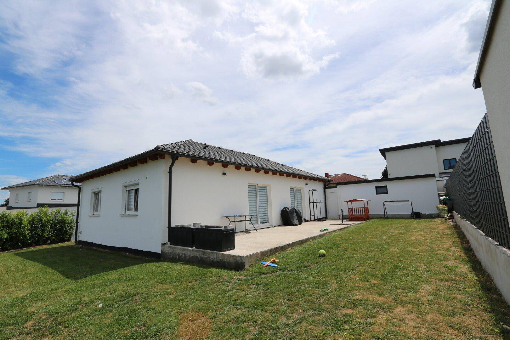 Rückseite des Hauses mit einer betonierten Terrasse, Gartenmöbeln und einer angrenzenden Rasenfläche.