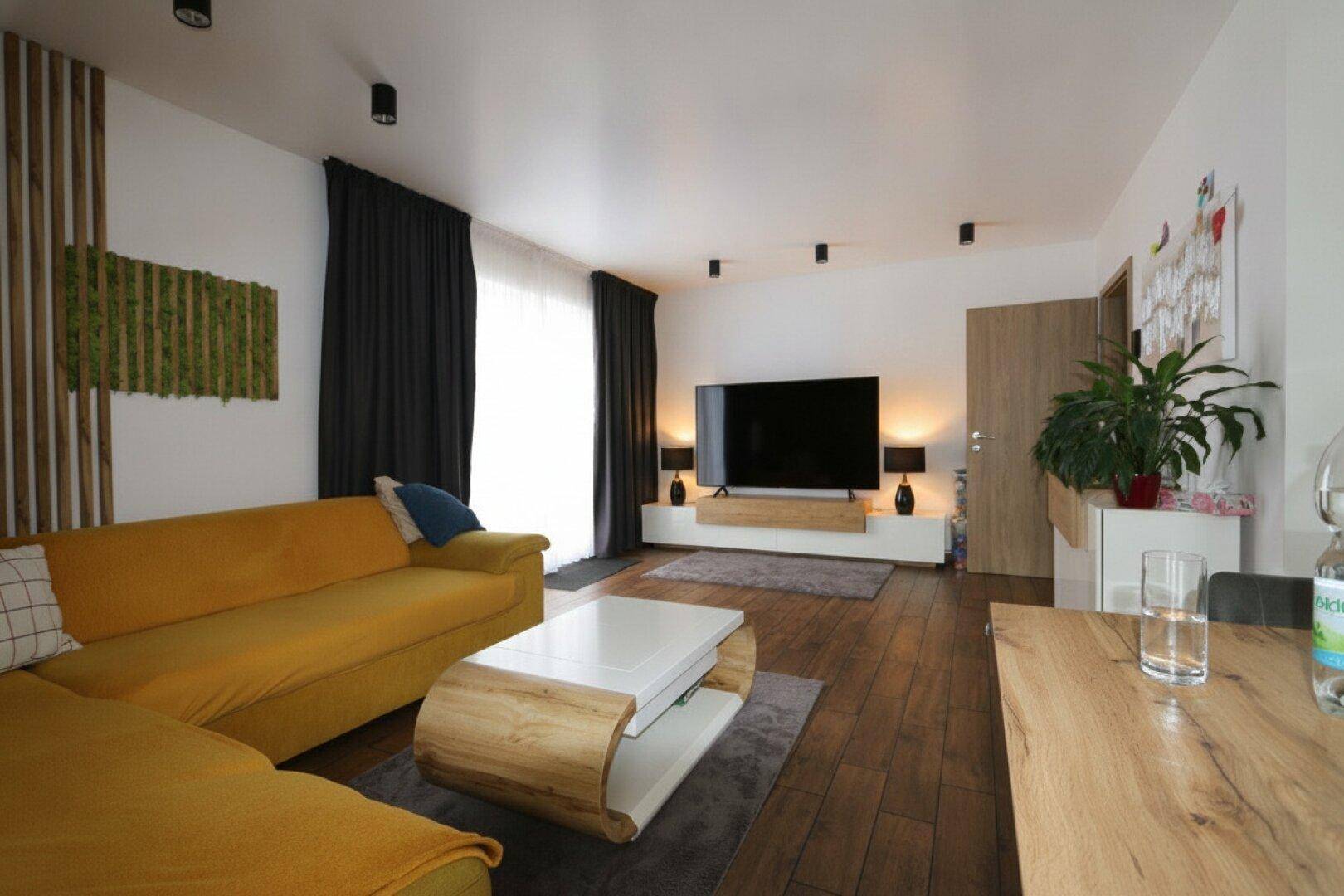 Geräumiges Wohnzimmer mit großem Fernseher, gelbem Sofa und Holzboden.