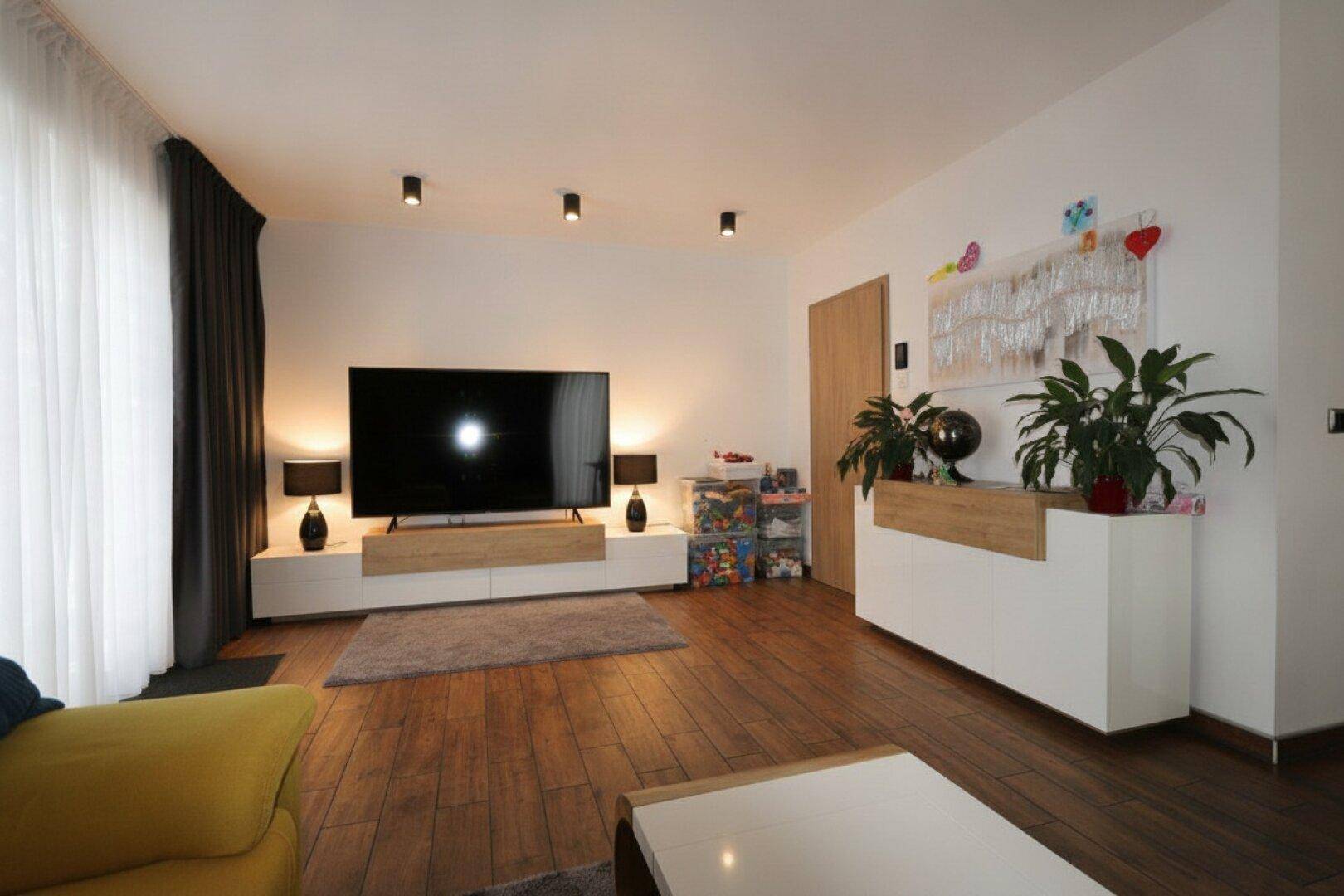 Geräumiges Wohnzimmer mit großem Fernseher, modernen Möbeln und Holzboden.