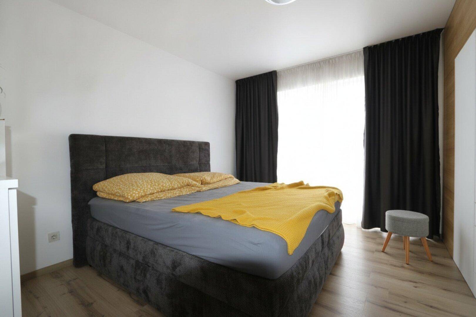 Schlafzimmer mit großem Boxspringbett, Holzboden und dunklen Vorhängen.