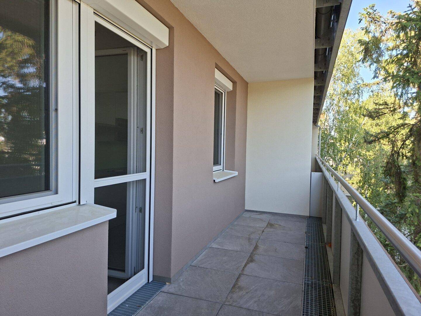 Schmaler Balkon mit Fliesenboden, Metallgeländer und Zugang durch große Fenster mit Rollläden.