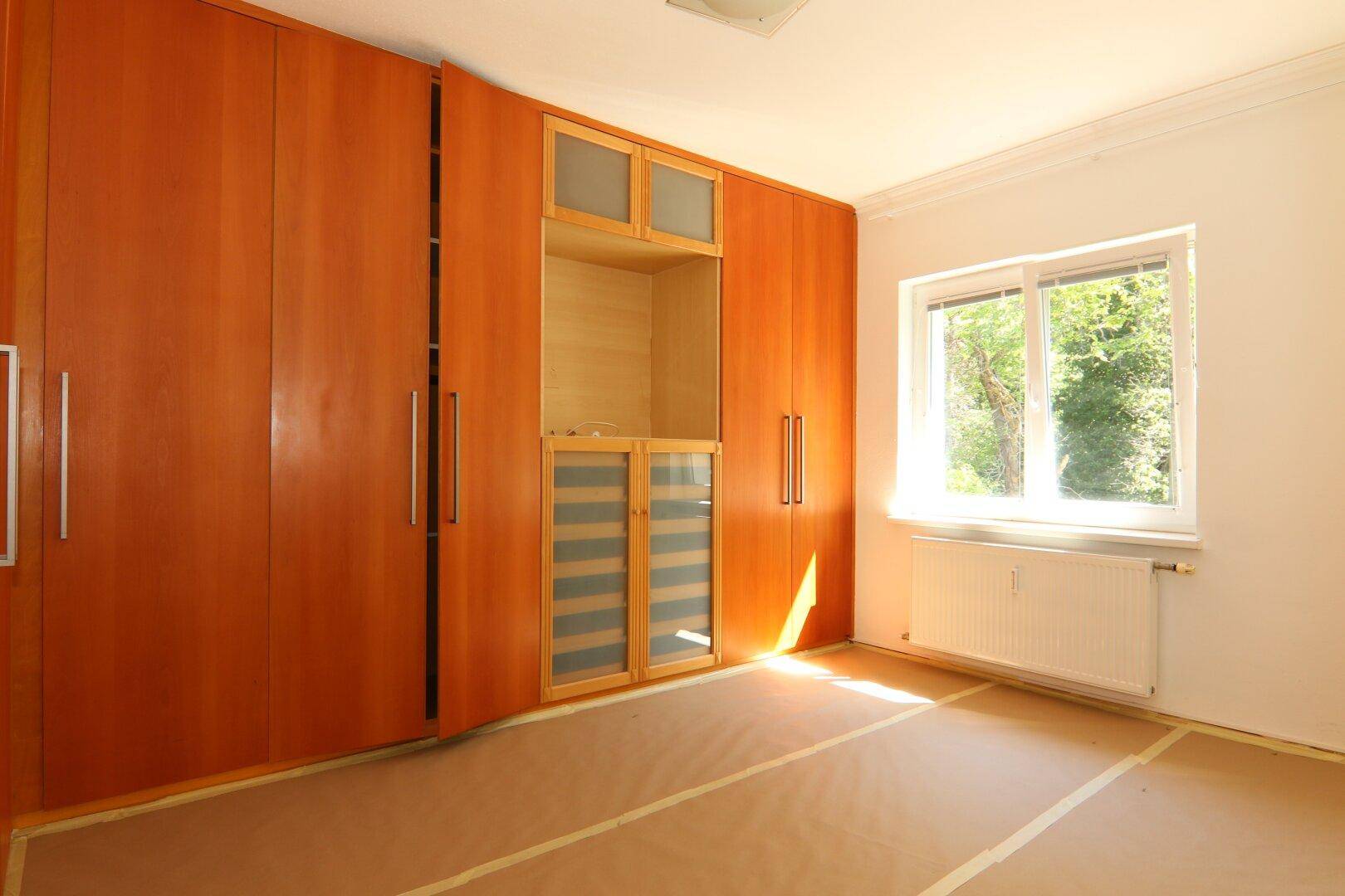 Helles Zimmer mit umfangreichen Holzeinbauschränken und Blick ins Grüne durch das Fenster.