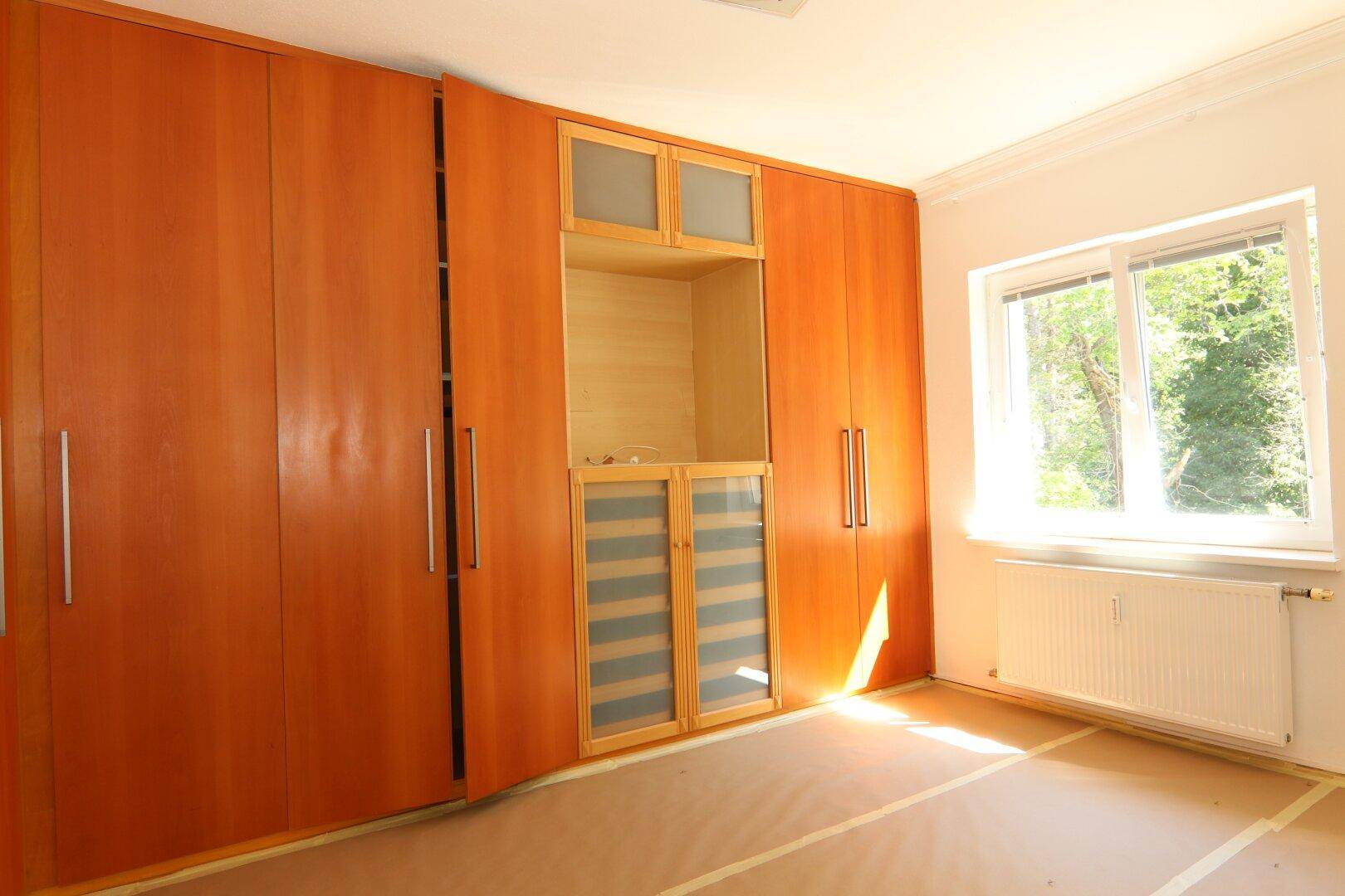 Helles Zimmer mit umfangreichen Holzeinbauschränken und Blick ins Grüne durch das Fenster.