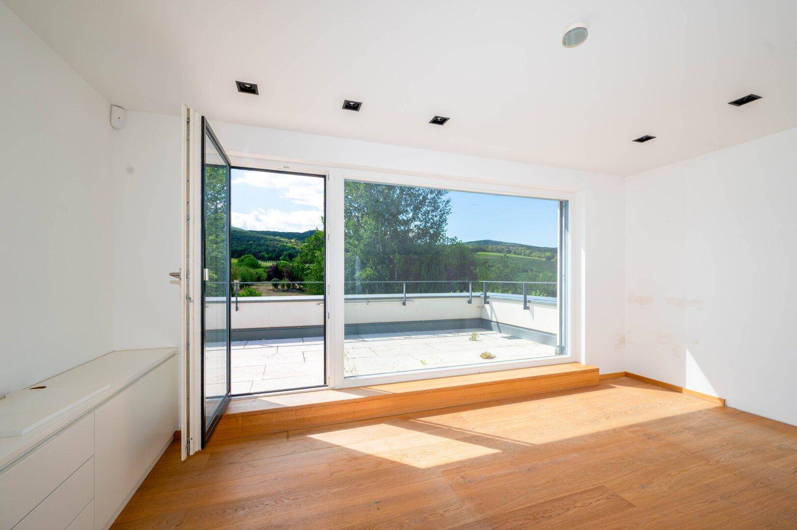 Heller Raum mit Holzboden und Zugang zu einer Dachterrasse mit weitem Blick auf die Landschaft.