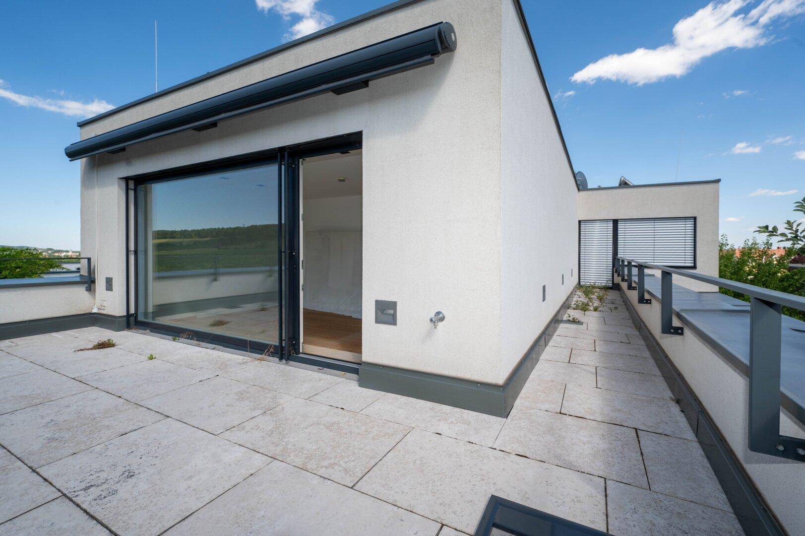 Weitläufige Dachterrasse mit Fliesenboden und Panoramablick auf die grüne Umgebung unter blauem Himmel.