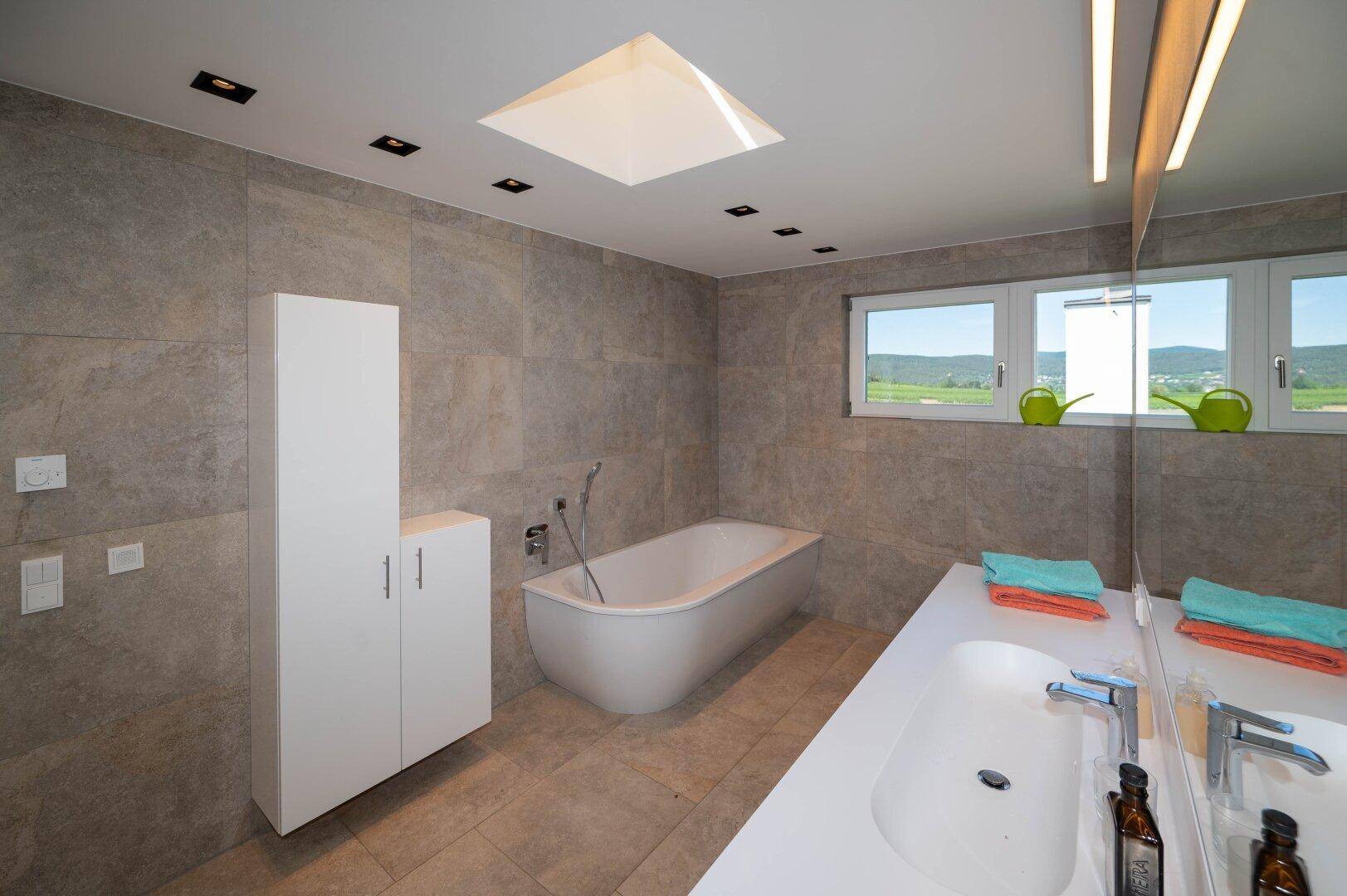 Helles Badezimmer mit Badewanne, großem Spiegel und Fenster mit Blick ins Grüne.