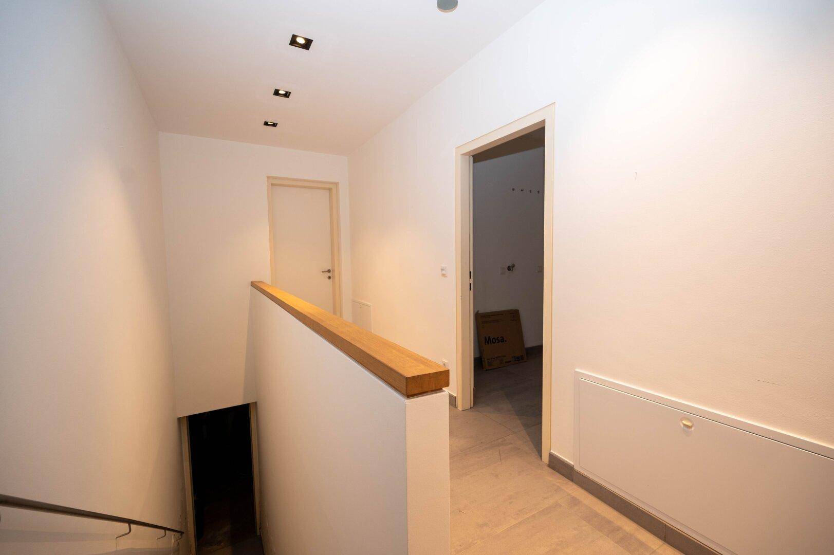 Heller Flur mit Treppenabgang und schlichten weißen Wänden, modern beleuchtet.