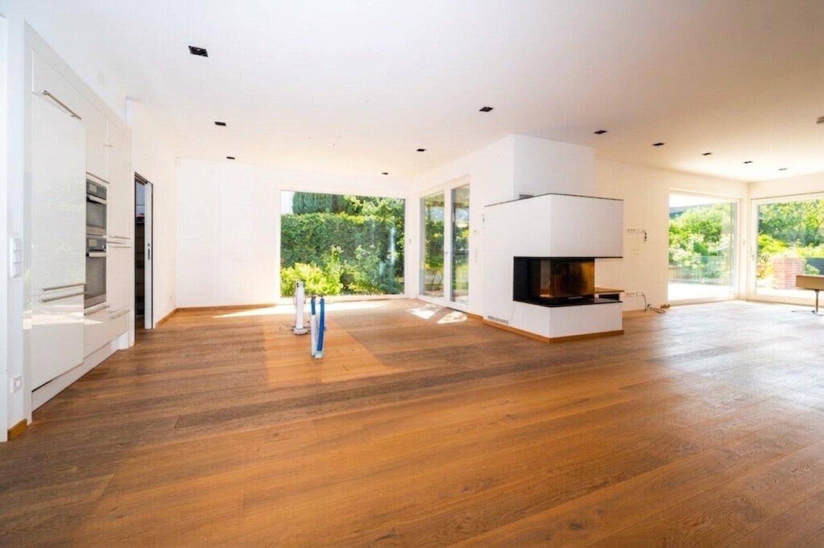 Helles Wohnzimmer mit Kamin, Holzboden und großen Fenstern, die viel Tageslicht hereinlassen.