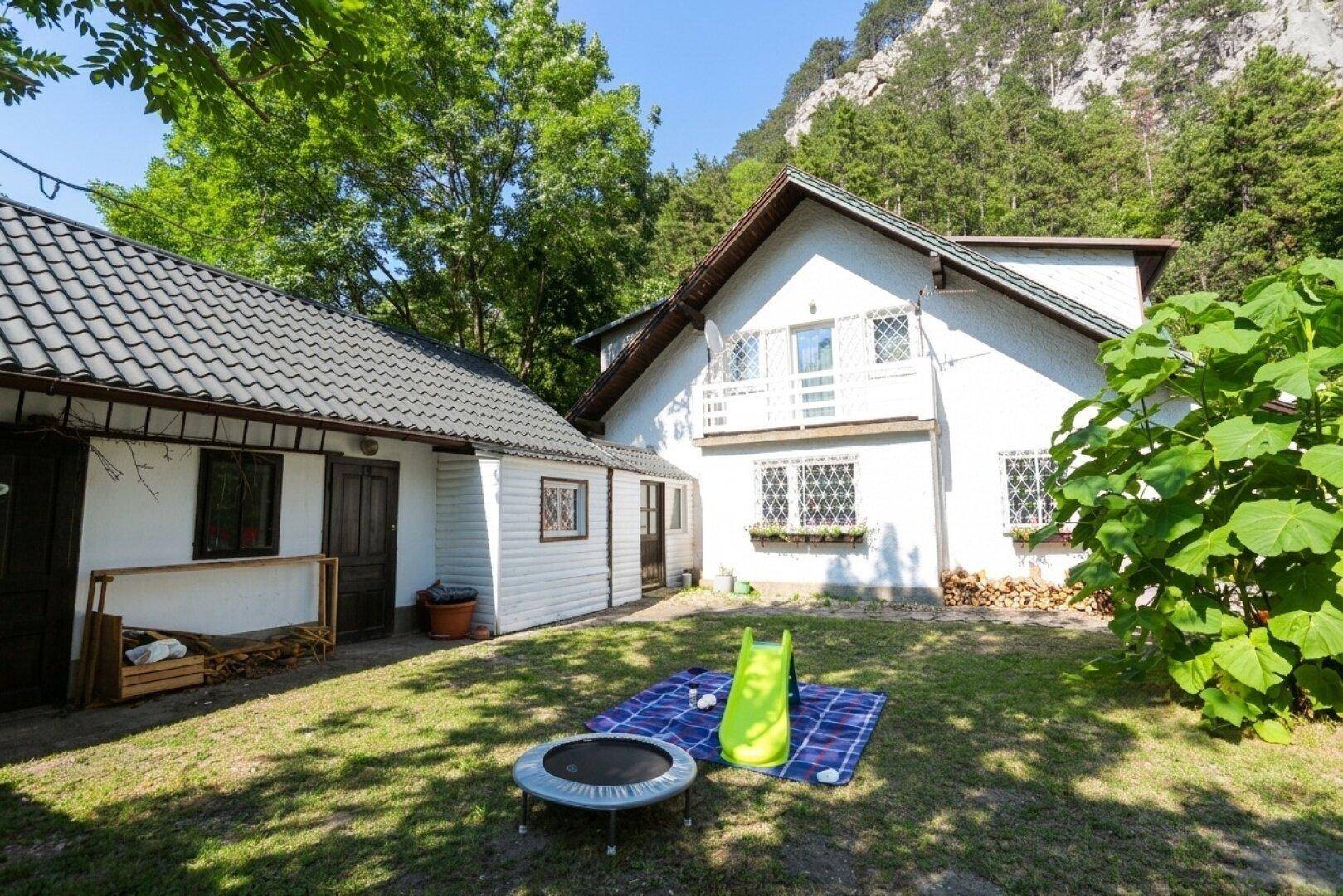 Rückansicht des Hauses mit weitläufigem Garten, Spielgeräten und Blick auf die bewaldeten Hügel.