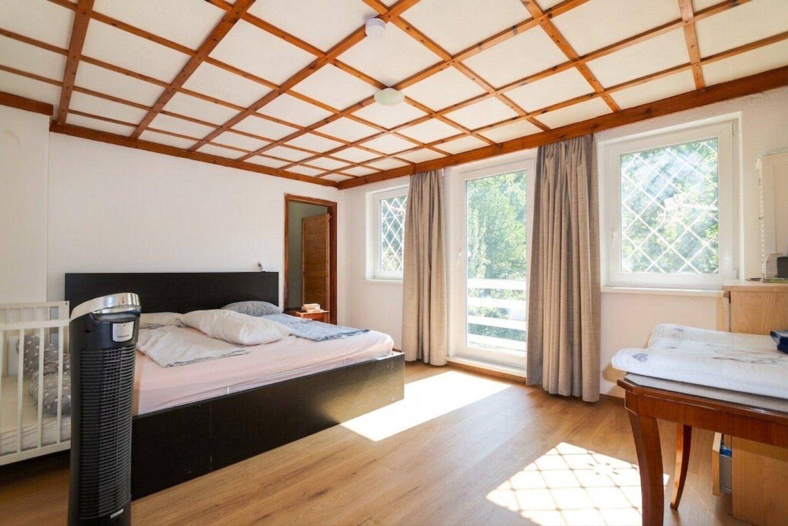 Helles Schlafzimmer mit Doppelbett, Babybett und Zugang zum Balkon, Holzbalkendecke.