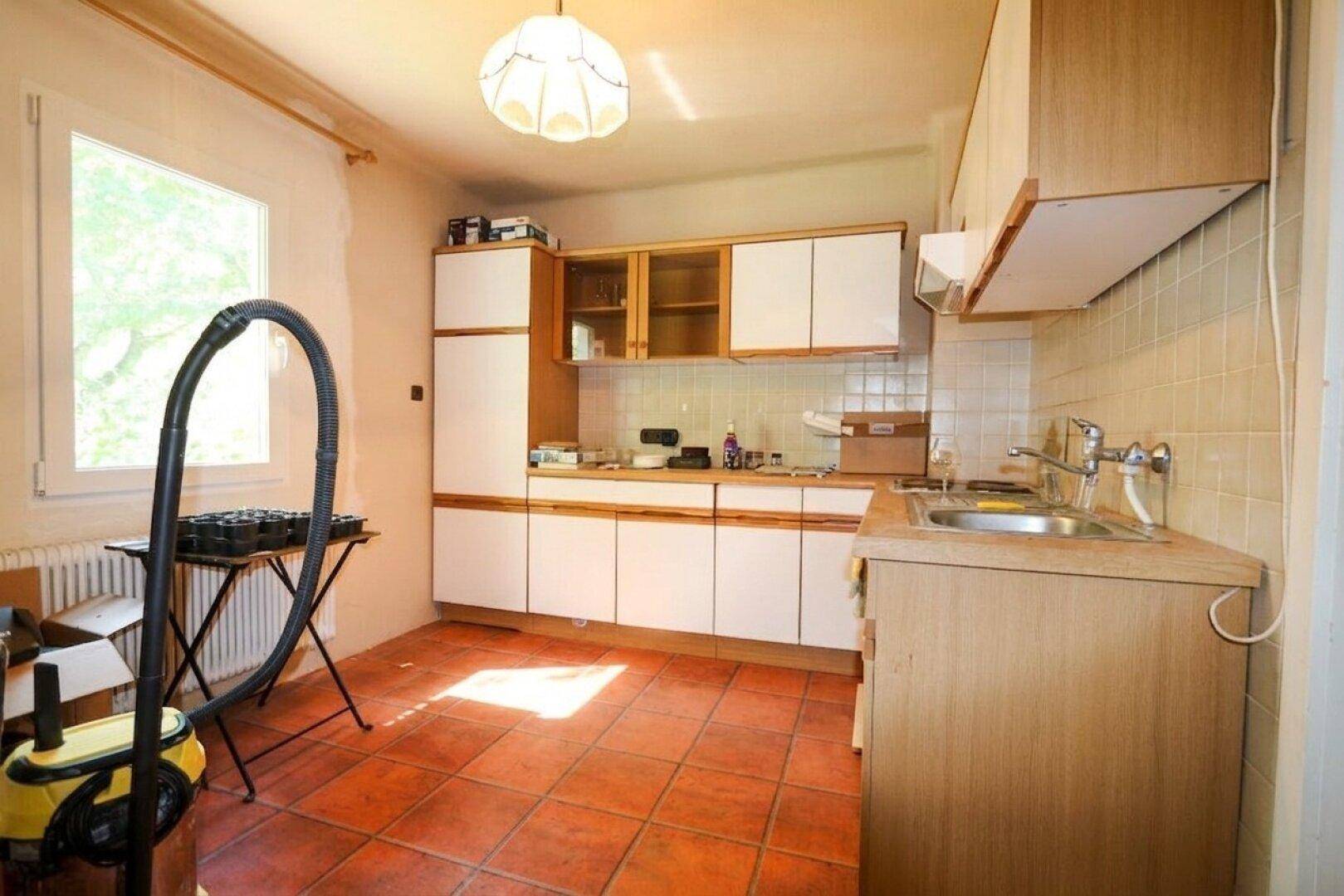 Ältere Küche mit Holzschränken, Terrakotta-Fliesenboden und Fenster.