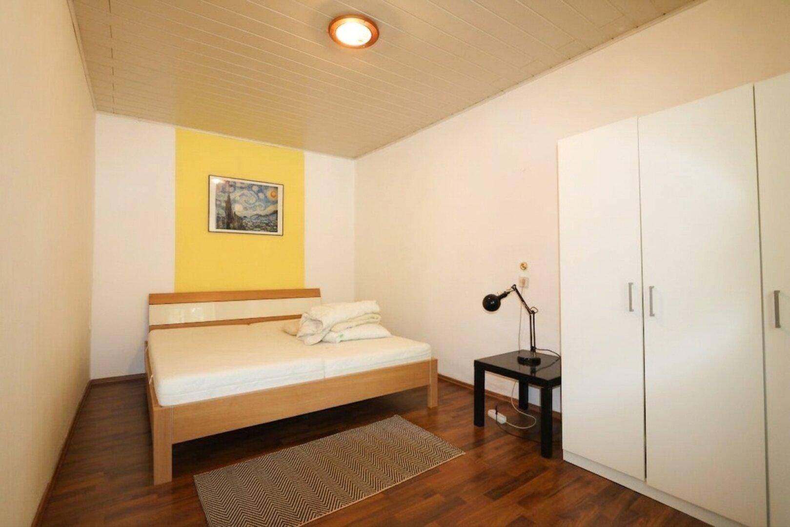 Schlafzimmer mit Holzbett, weißem Kleiderschrank, Nachttisch und gelber Akzentwand.