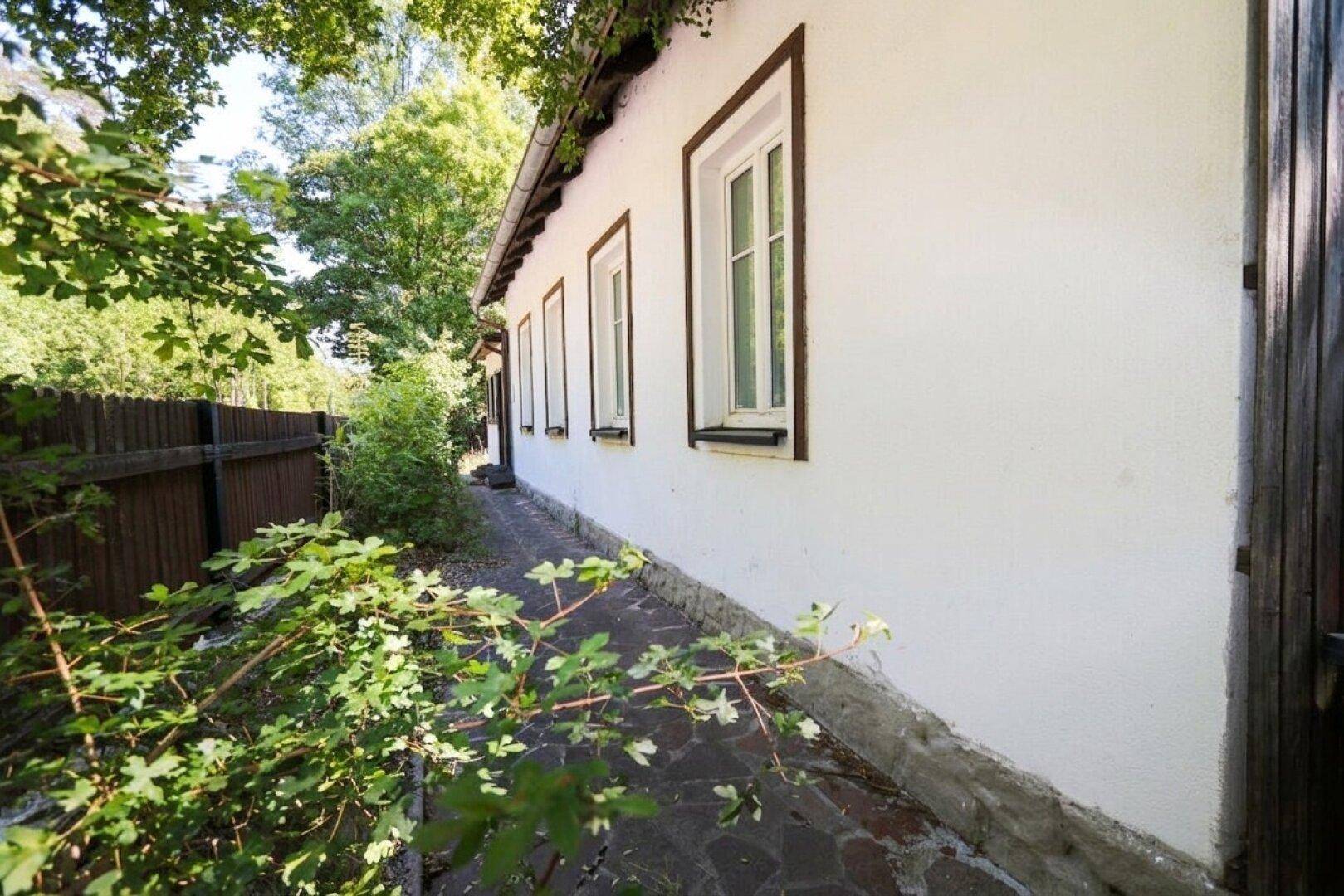 Weiße Hausfassade mit dunklen Fensterrahmen, umgeben von einem Steinweg und viel Grün.