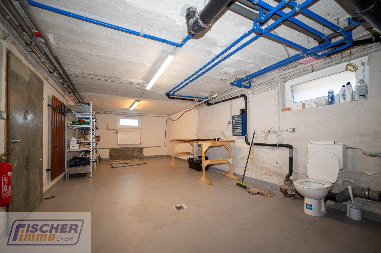 Geräumiger Kellerraum mit sichtbaren Rohren, Toilette und einfacher Werkbank.