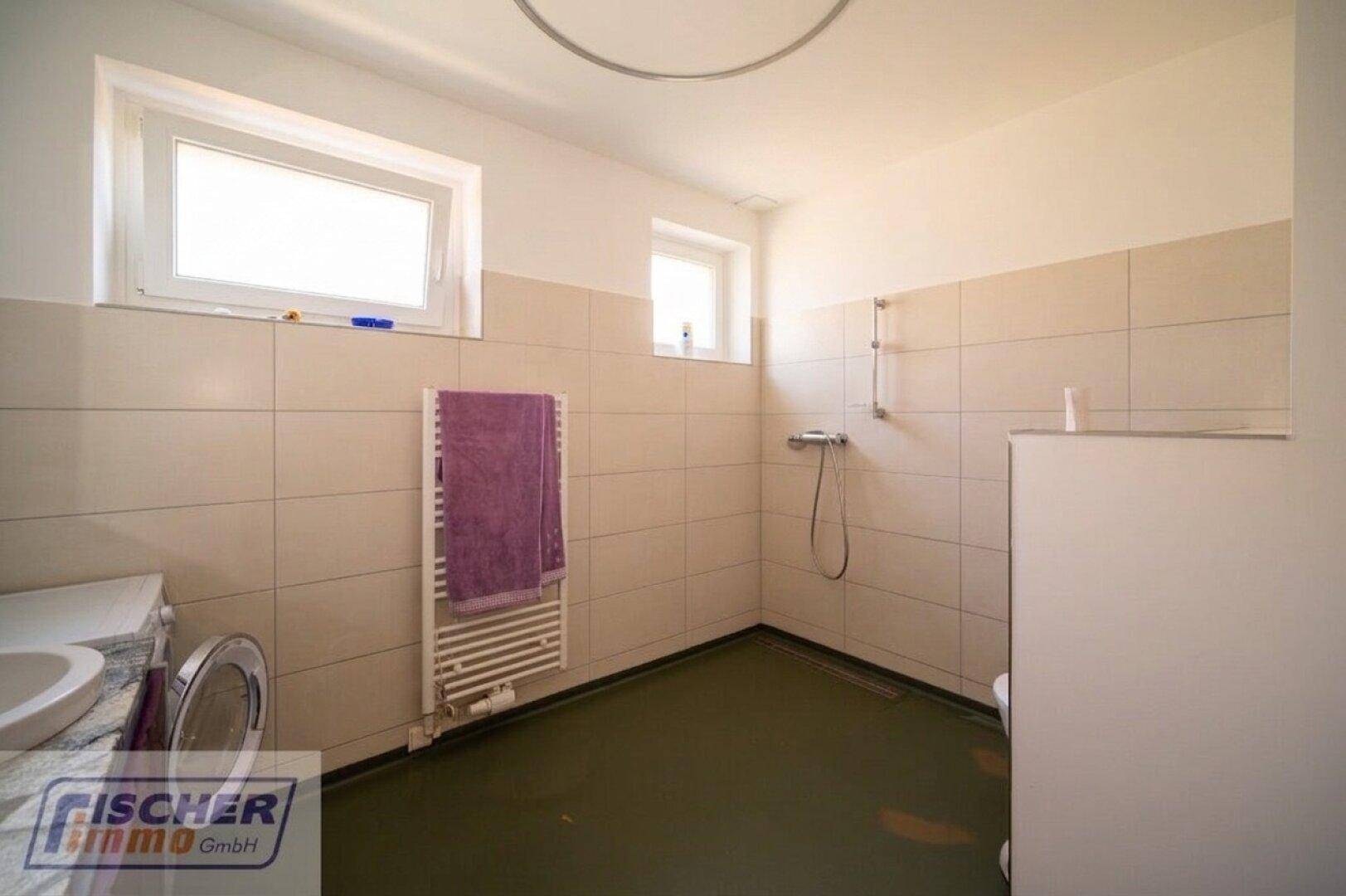 Badezimmer mit beigefarbenen Fliesen, Dusche und Fenster für Tageslicht.