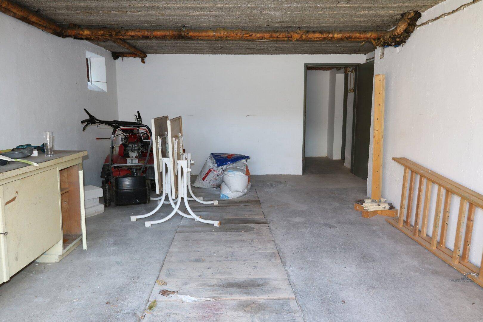 Unfertiger Kellerraum mit Betonboden, freiliegenden Rohren und verschiedenen Lagergegenständen.