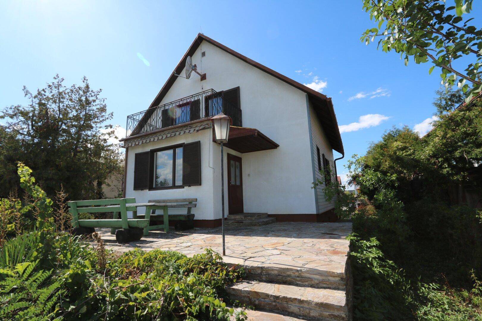 Hausansicht mit Steinterrasse, Gartenbereich und einem kleinen Balkon unter blauem Himmel.