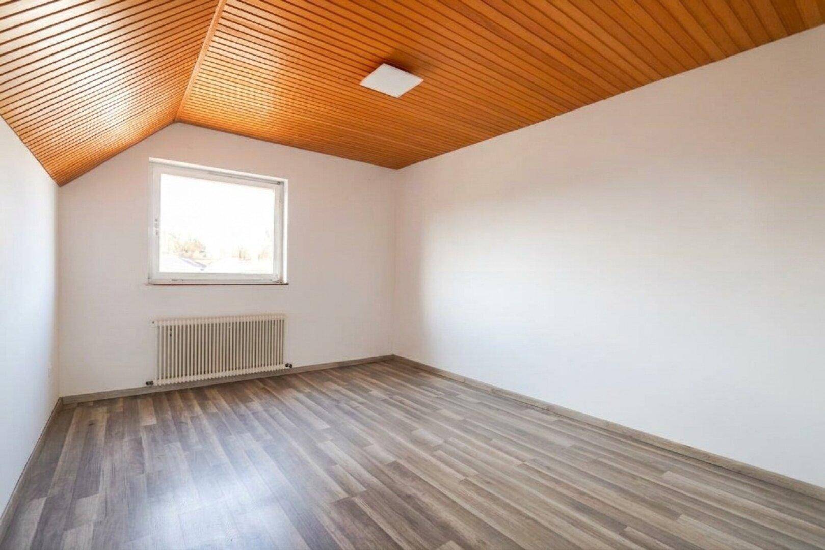 Helles Schlafzimmer im Dachgeschoss mit Laminatboden, Holzdecke und einem Fenster.