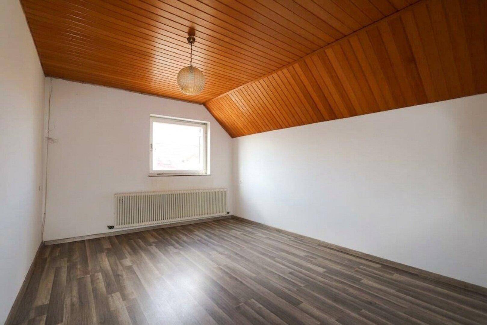 Helles Zimmer im Dachgeschoss mit Holzdecke, Laminatboden, Fenster und Heizkörper.