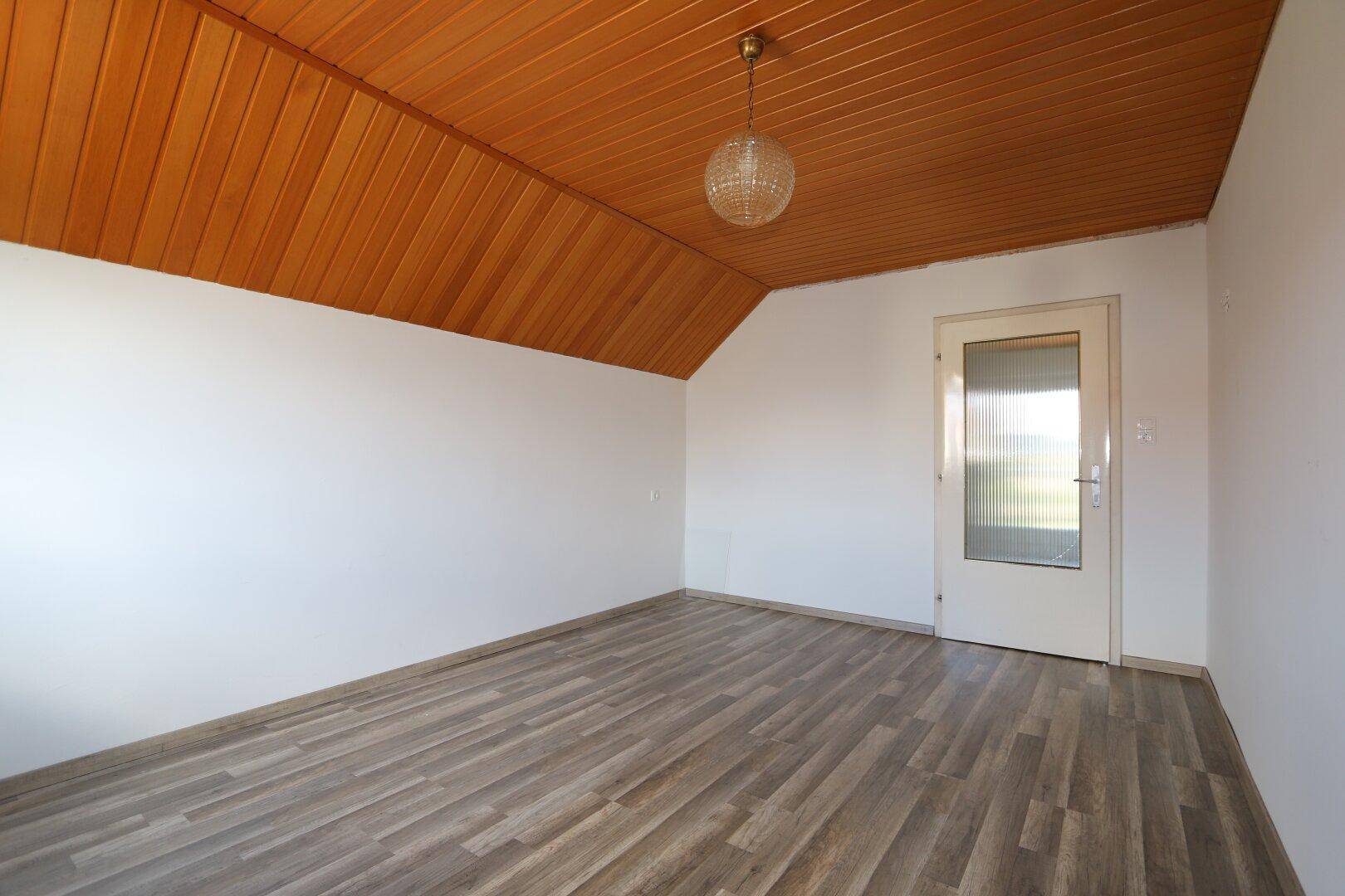 Geräumiges Zimmer mit Holzpaneeldecke, Laminatboden und einer Tür mit Glaseinsatz.