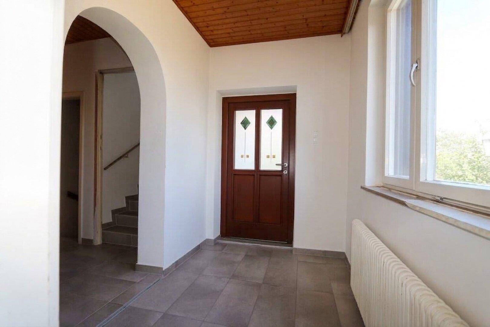 Heller Eingangsbereich mit Fliesenboden, Holzdecke und einer braunen Eingangstür mit Glaseinsätzen.