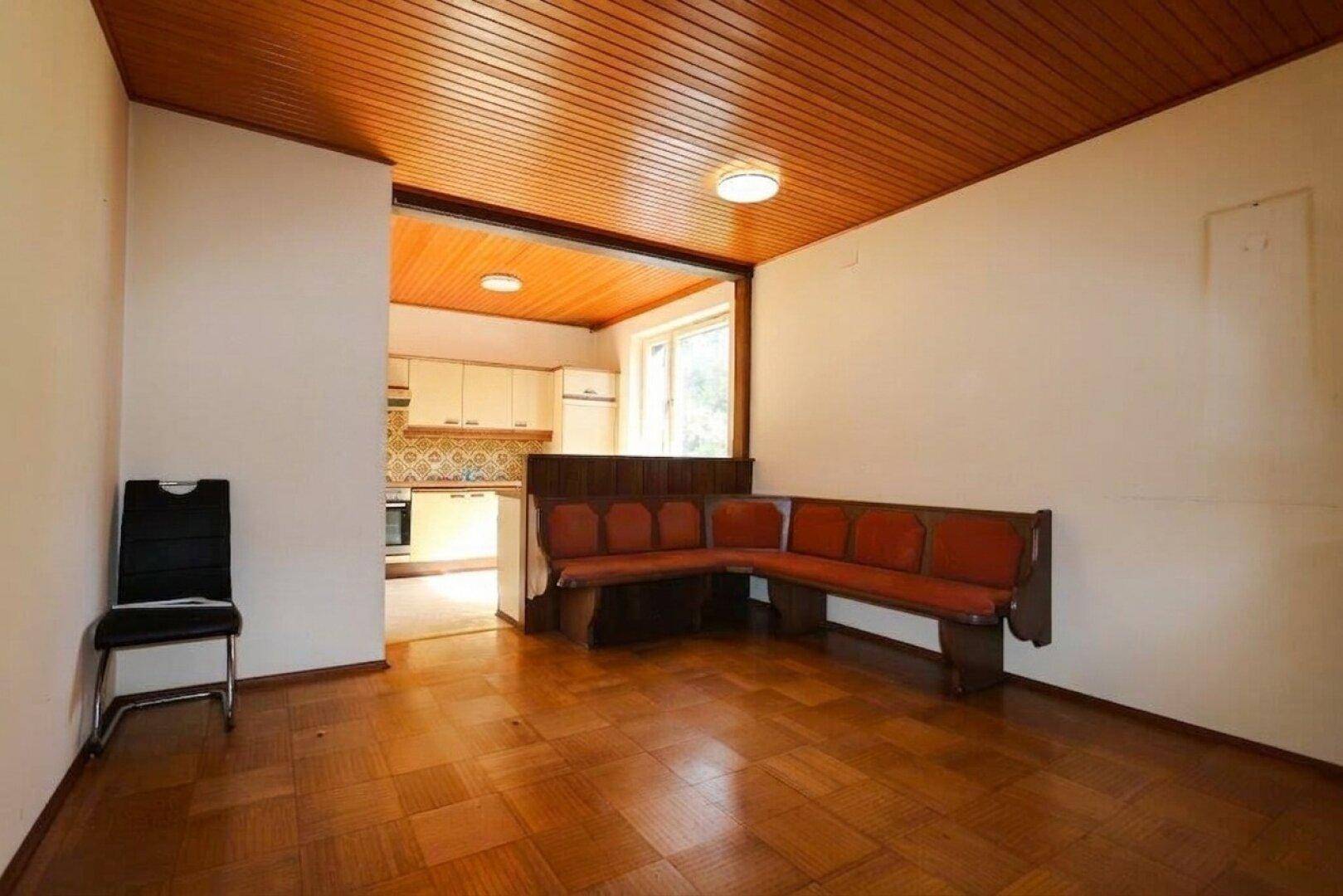 Wohnbereich mit Parkettboden, Holzdecke und einer traditionellen Eckbank, offen zur Küche.