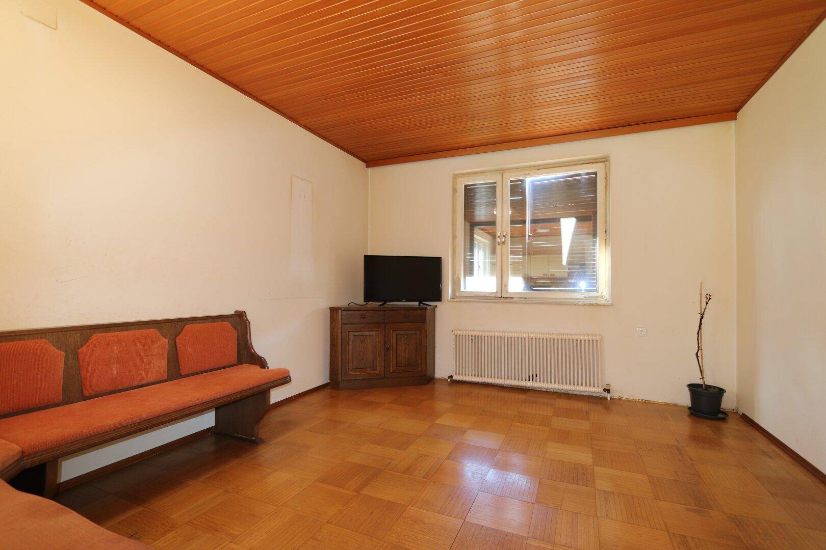 Gemütliches Wohnzimmer mit Parkettboden, Holzdecke und einem Fenster mit Blick ins Grüne.