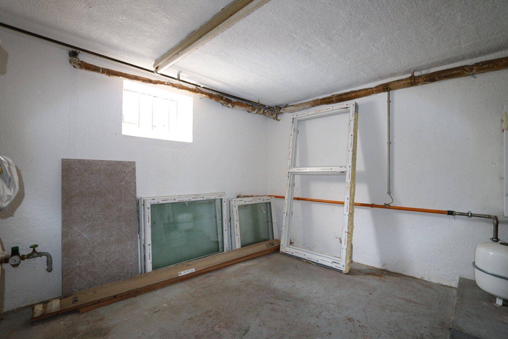 Kellerraum mit Betonboden, kleinen Fenstern, sichtbaren Installationen und gelagerten Fenstern.