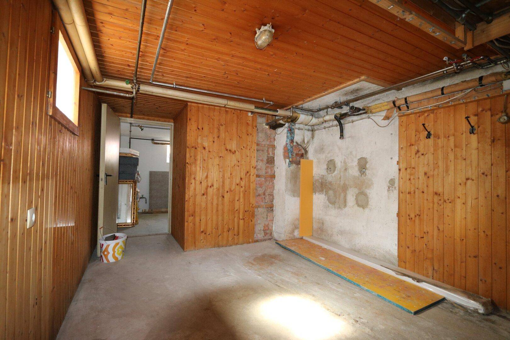 Rustikaler Kellerraum mit Holzpaneelen, Betonboden, sichtbaren Rohren und natürlichem Lichteinfall.