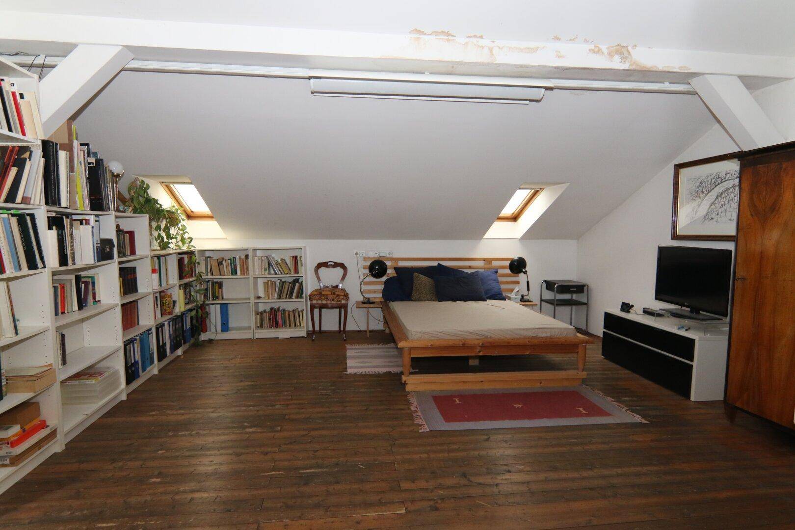 Schlafzimmer in einer Dachgeschosswohnung mit Holzboden, Bücherregalen und Dachfenstern für natürliches Licht.