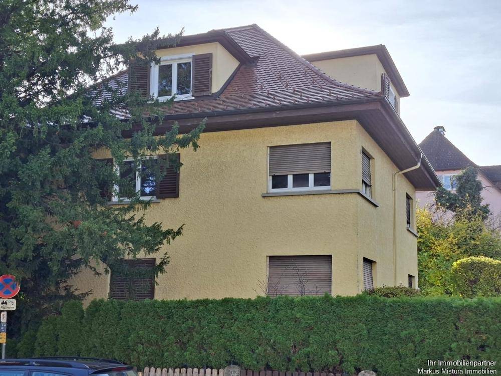 Gelbes Mehrfamilienhaus mit braunen Fensterläden und einem Satteldach, umgeben von Bäumen.