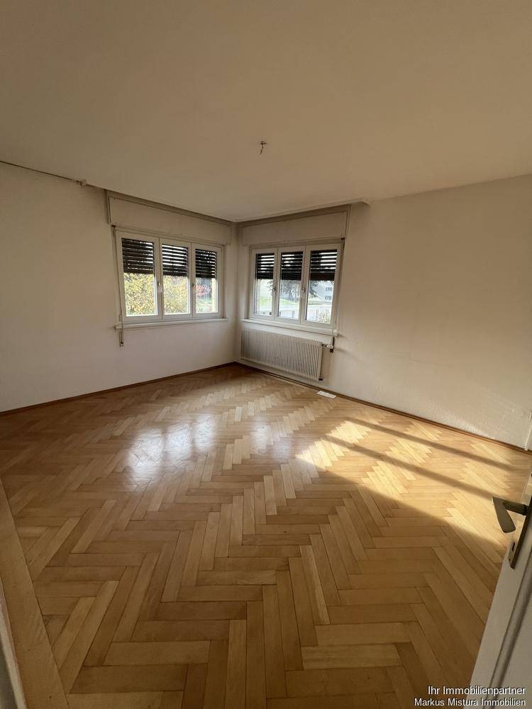 Helles Zimmer mit Fischgrätparkett und großen Fenstern, ideal als Wohnzimmer.