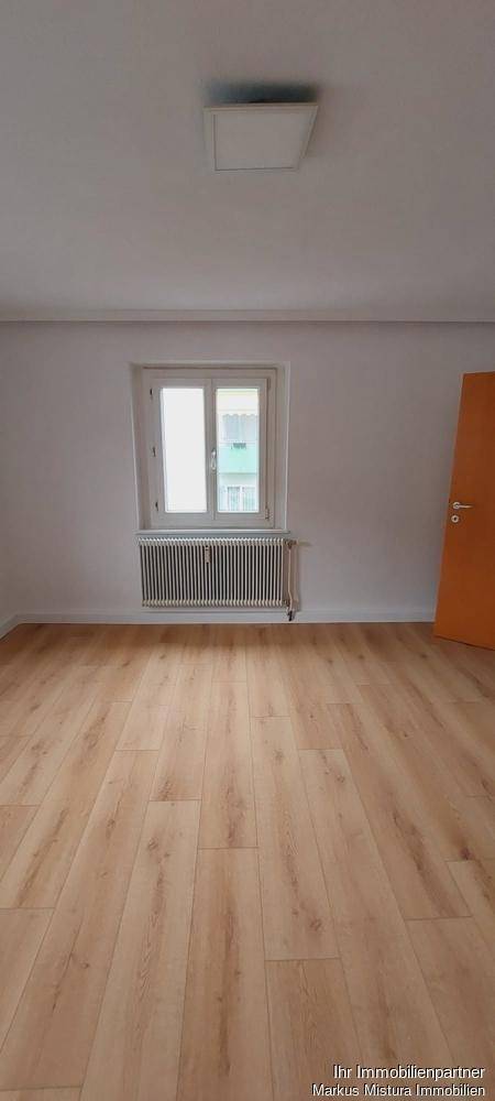 Leeres Zimmer mit hellem Laminatboden, weiß gestrichenen Wänden und einem Fenster mit Heizkörper.