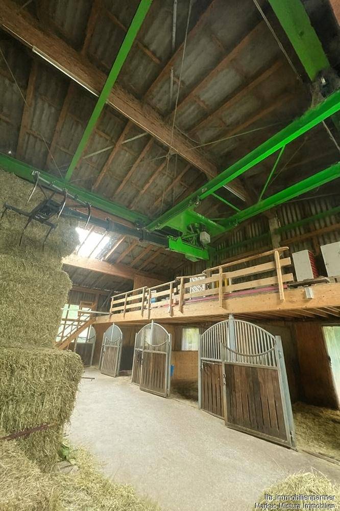 Innenansicht eines Stalls mit Heuballen, Pferdeboxen und einer Hebefunktion unter dem Dach.