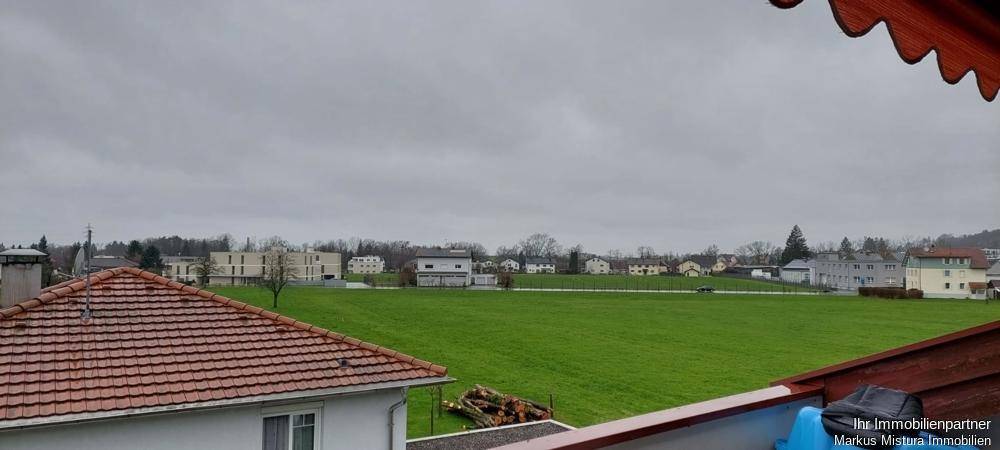 Weitblick über grüne Felder und Dächer von einem Balkon aus an einem bewölkten Tag.