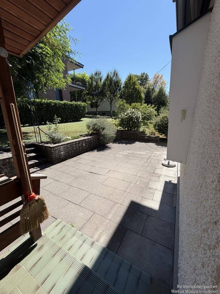 Gepflasterte Terrasse neben dem Haus, umgeben von Grünflächen und mit Zugang über Stufen.