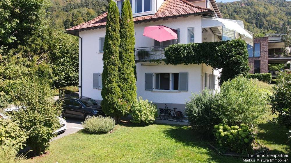 Seitenansicht des Hauses mit gepflegtem Rasen, hohen Bäumen und einem Balkon mit Sonnenschirm.