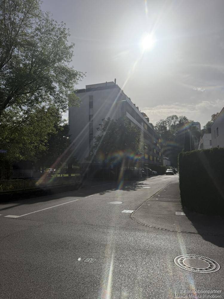 Gebäudeansicht von der Straße mit viel Sonnenlicht und Bäumen im Vordergrund.