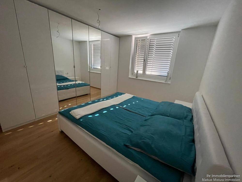 Schlafzimmer mit Doppelbett, großem Spiegelschrank, hellem Holzfußboden und Fenster mit Jalousien für Privatsphäre.