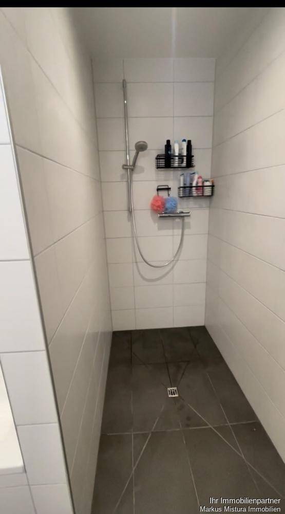 Geräumige Duschkabine mit weißen Fliesen, Duschkopf und praktischer Ablage für Pflegeprodukte, barrierefrei gestaltet.