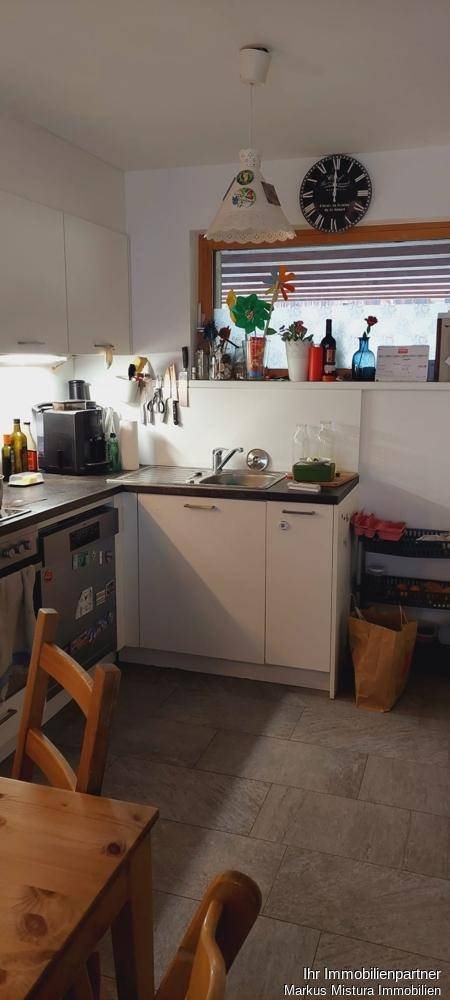 Helle Küche mit weißen Einbauschränken, Spülmaschine und einem Esstisch aus Holz am Fenster.