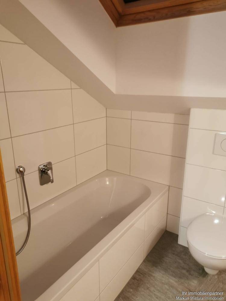 Dachgeschoss-Badezimmer mit Badewanne und Toilette, umgeben von hellen Fliesen und schräger Decke.