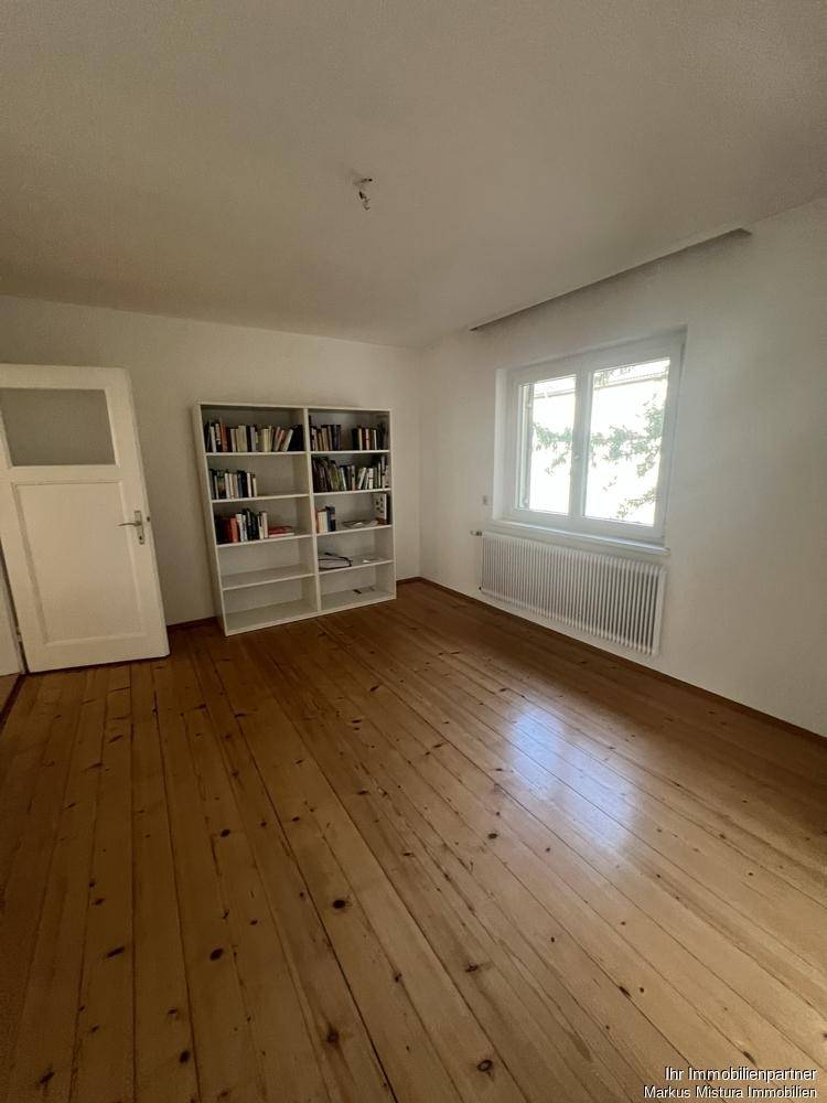 Helles Zimmer mit Holzdielenboden, großem Fenster und einem weißen Bücherregal an der Wand.