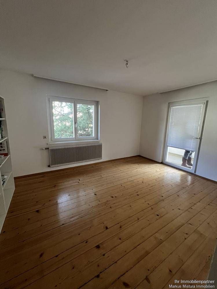 Leeres Zimmer mit Holzdielenboden, großem Fenster und Zugang zu einem Balkon.