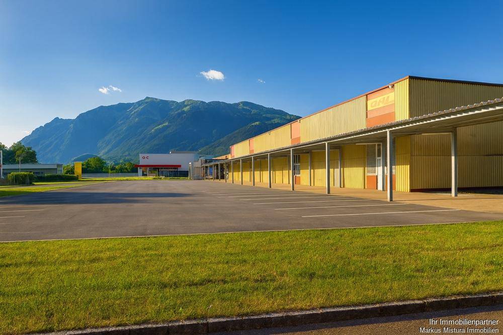 Weitläufiger Parkplatz vor einem Industriegebäude mit grüner Rasenfläche und beeindruckendem Bergblick.