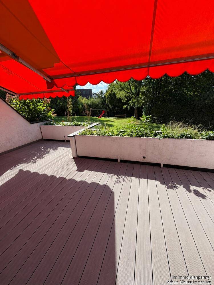 Große Holzterrasse mit ausgefahrener roter Markise und Blick ins Grüne.