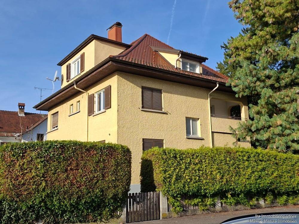 Ansicht eines gelben Mehrfamilienhauses mit rotem Ziegeldach und grüner Hecke im Vorgarten.