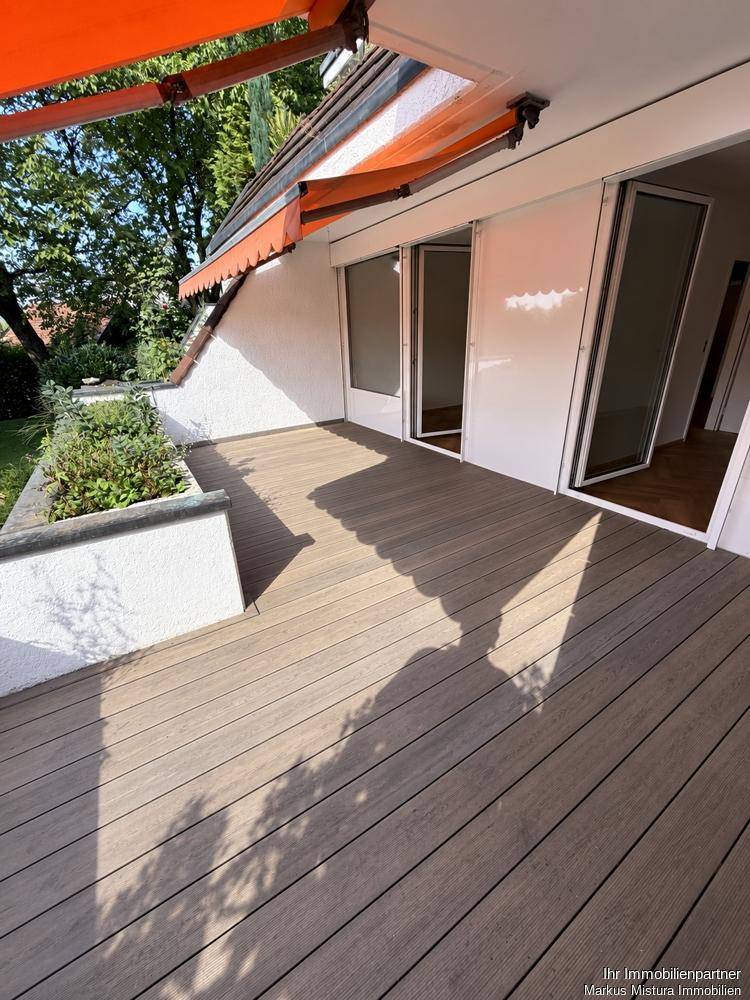 Moderne Holzterrasse vor der Fassade mit großen Glasfronten und viel Sonnenlicht.