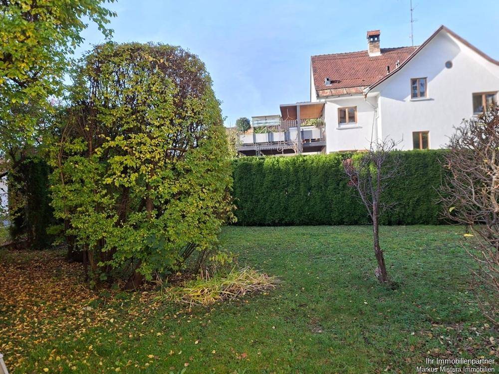 Gepflegter Garten mit Rasenfläche, hohen Hecken und Bäumen, umgeben von weiteren Häusern.