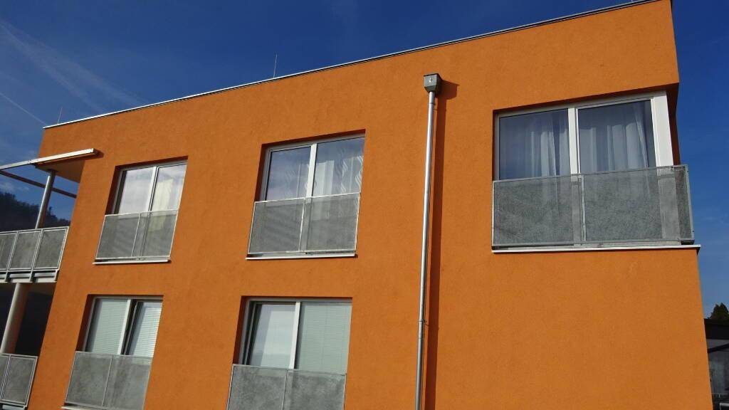Moderne Fassade eines Neubaus mit leuchtend orangefarbenem Putz und französischen Balkonen unter blauem Himmel.